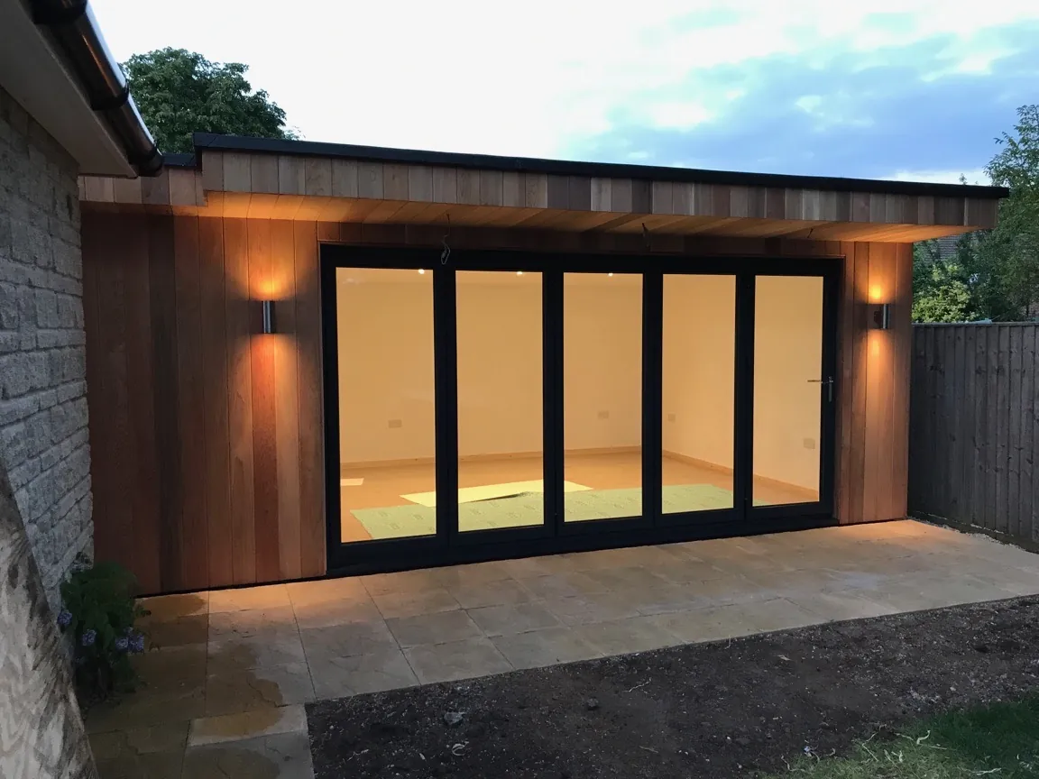 A cedar-clad garden room lit up in the evening, its bi-folding doors closed